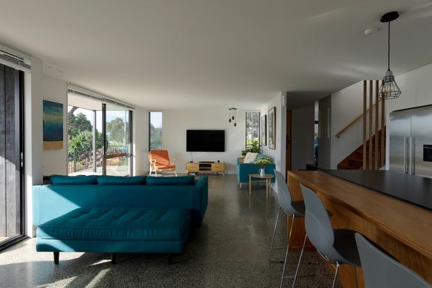 Open-plan contemporary living area and kitchen with a large teal sectional sofa, wood media console, wall-mounted television, dining or kitchen island with timber cabinetry and dark benchtop, and grey bar stools. Full-height glazing opens toward a balcony and leafy outlook, while a timber stair with a simple railing leads upward. The room has polished concrete or terrazzo-like flooring, white walls and ceiling, abundant daylight, and pendant lighting.