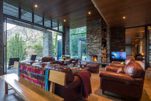 A spacious rustic-modern living room with floor-to-ceiling glazing overlooking a dramatic rocky mountain landscape. The room features exposed stacked-stone walls and fireplace, a warm timber slatted ceiling, timber flooring, extensive dark-framed glazing, and multiple brown leather sofas and armchairs arranged around a central fireplace and television. Colourful woven throws add accents to the otherwise earthy interior.