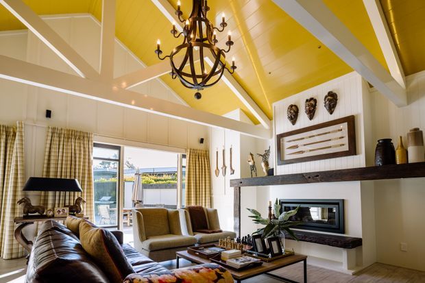 A spacious, warm living room with a vaulted yellow-painted timber ceiling, exposed white beams, white vertical wall panelling, and large windows dressed with checked curtains. The room features a dark leather sofa, upholstered armchairs, a substantial timber mantel shelf, fireplace, framed artwork, sculptural wall decor, and an ornate dark metal chandelier. Outdoor scenery is visible through the glazing.