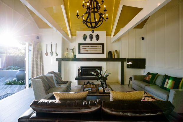 A richly styled living room with a large dark leather sofa in the foreground, two upholstered armchairs, a green upholstered sofa, and a central coffee table. The room features white paneled walls, a dramatic mustard-yellow and white coffered ceiling, a dark fireplace mantel with framed artwork above it, and a black globe chandelier. Glazed doors at left open to an outdoor deck or terrace, bringing in abundant natural light.