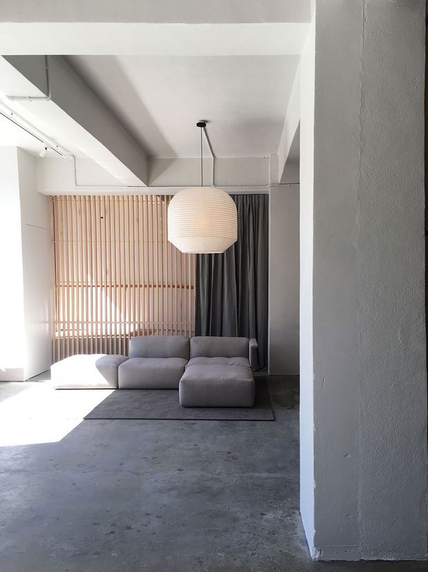 Minimalist living room with a low modular grey sectional sofa on a dark rug, a large rounded paper pendant light, and a warm timber slatted screen behind the seating. Dark full-height curtains, exposed concrete flooring, white painted structural beams and columns, and strong natural light create a restrained loft-like interior.