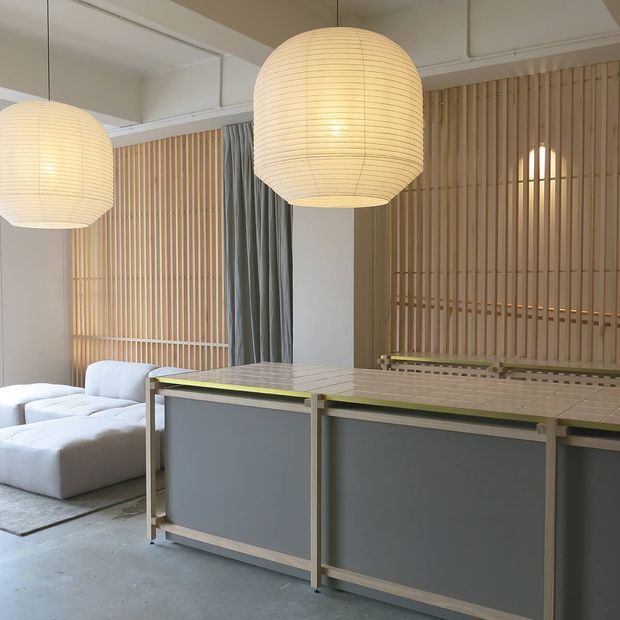 A softly lit contemporary interior featuring a large tiled island or counter in the foreground, pale timber vertical slatted wall screens, oversized woven pendant lights, a grey curtain, and a white sectional sofa. The space has a restrained neutral palette with warm timber detailing and a polished concrete floor.