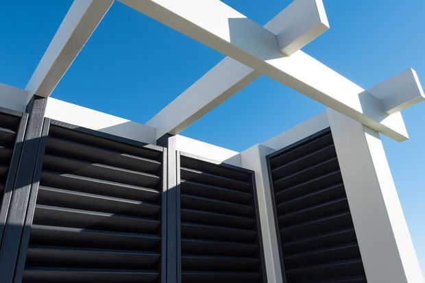 Upward-looking architectural detail of a modern exterior courtyard or screening structure, featuring dark horizontal louvred panels framed by crisp white geometric beams and posts against a clear blue sky.