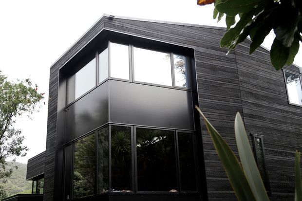Exterior view of a contemporary two-storey house with a strong cubic form, dark horizontal timber cladding, extensive black-framed glazing, and a projecting upper-level volume. The building sits in a leafy, sloped landscape and is partly screened by foreground planting.