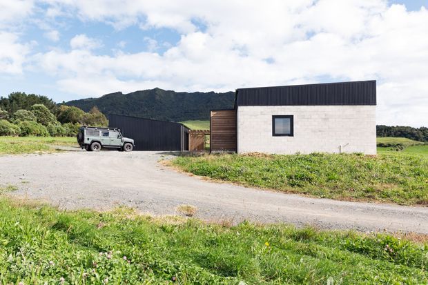 A rural contemporary building complex viewed from outside, featuring a simple white masonry block, a dark pitched metal roof, dark cladding, timber elements, and a gravel driveway. Rolling green pasture and a wooded hillside surround the property.