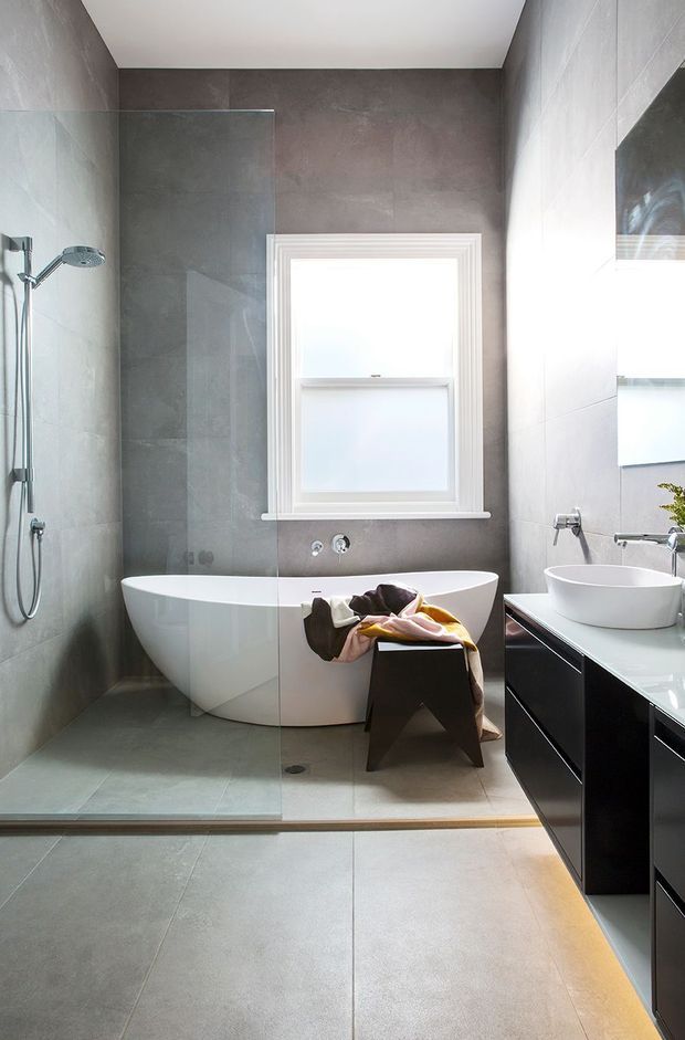 A contemporary bathroom with a walk-in glass shower, freestanding white bathtub, wall-mounted chrome shower fittings, vessel basin, and dark floating vanity cabinetry. Large grey tiles cover the walls and floor, with a bright frosted window and white trim providing natural light.