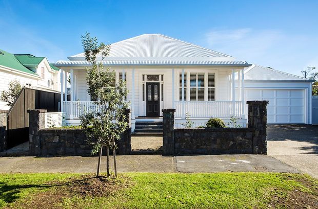 Street-facing exterior of a single-storey white weatherboard house with a broad hipped corrugated metal roof, front veranda with white balustrade, traditional detailing, and an attached single garage. A low dark masonry boundary wall and front garden sit between the house and the pavement.