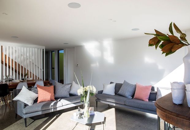 Bright contemporary living room with two grey sofas, pastel accent cushions, a white coffee table, and a rounded timber side table. A dining area and staircase are visible behind a vertical slatted divider, with strong natural light entering from the right.
