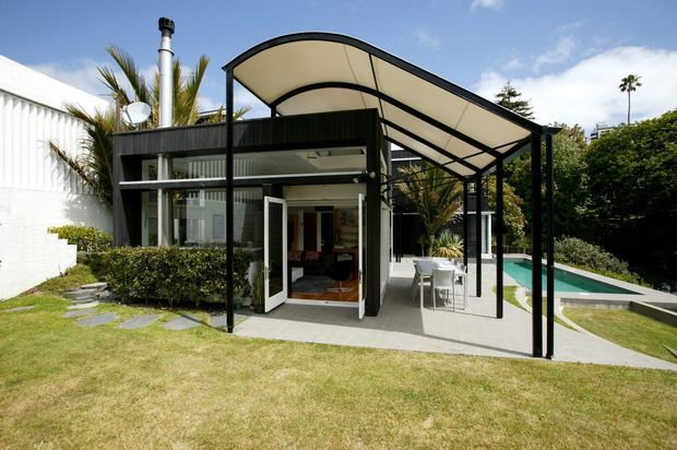 Exterior view of a contemporary residential pavilion or extension with extensive glazing, dark structural framing, and a large curved translucent canopy over a paved outdoor entertaining area. An open living space is visible inside, with a dining setting beneath the canopy and a swimming pool extending alongside the building. The property is surrounded by landscaped lawn, hedges, palms, and mature trees.