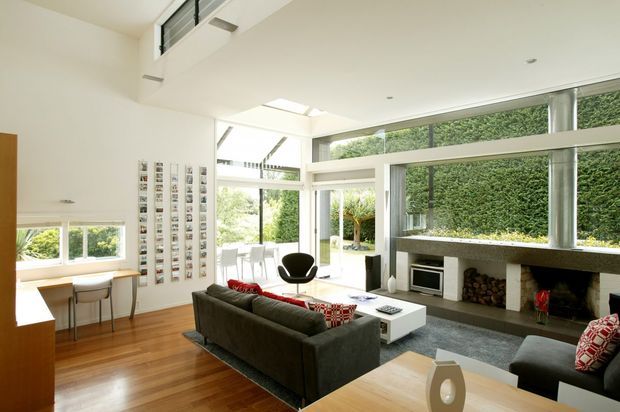 Bright open-plan living room with a contemporary architectural design. The space features expansive floor-to-ceiling glazing, a high ceiling with clerestory windows and skylights, polished timber flooring, a large dark sectional sofa, a white low coffee table, and a substantial stone or concrete fireplace wall with integrated shelving and stacked firewood. A compact dining or study nook is visible at the left, while a landscaped courtyard is seen through the glazing.