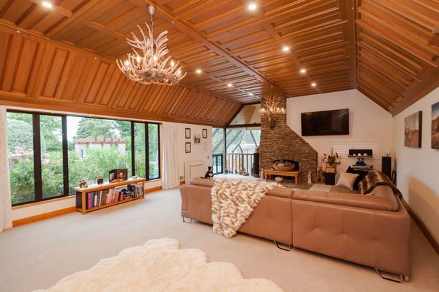 Spacious, warm living room with a dramatic pitched timber-lined ceiling featuring exposed beams and recessed lighting. The room includes a large tan sectional sofa, stone-clad fireplace with television above, chandelier lighting, pale carpet, framed artwork, and extensive black-framed glazing overlooking greenery.