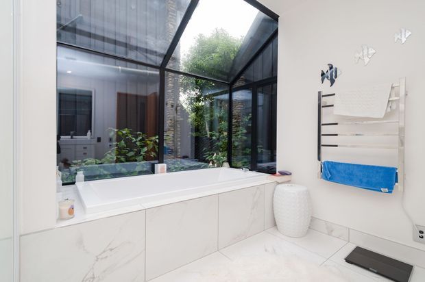 Bright contemporary bathroom featuring a built-in white bathtub beneath large black-framed glazed panels, white marble-look wall and floor finishes, and a wall-mounted towel ladder with a blue towel. A vanity area and lush courtyard greenery are visible through the glazing and reflections.