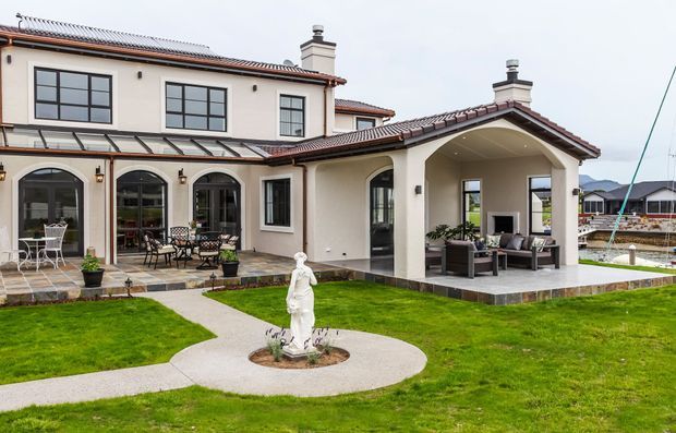 A large two-storey stucco residence with a red-brown tiled pitched roof, dark-framed windows, and multiple arched openings. The rear facade opens onto a paved patio and covered outdoor lounge, with manicured lawns, curved concrete paths, planter pots, and a central classical statue fountain.