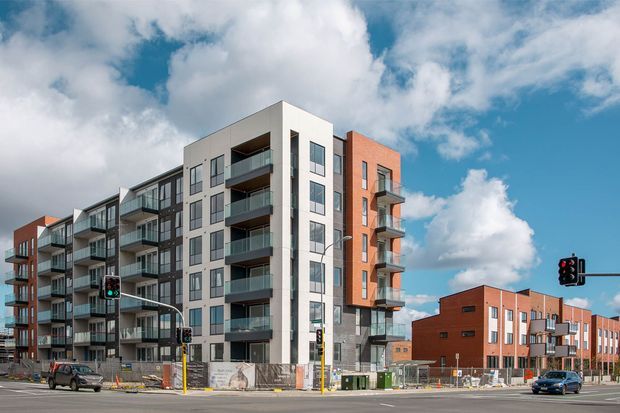 Exterior street view of a contemporary multi-storey apartment development with a mix of white rendered walls, dark framing, glazed balconies, and red-brick facade sections. The building sits on an urban corner beside a broad road intersection, with additional brick apartment buildings visible in the background and construction activity at street level.