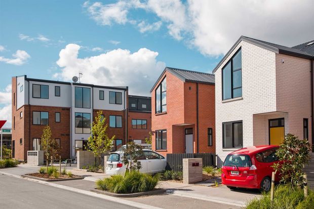 A sunny streetscape showing a contemporary multi-home residential development with attached and semi-detached brick-clad townhouses. The homes feature varied red, brown, and white brick facades, dark metal pitched roofs, large rectangular windows, compact landscaped front gardens, driveways, and parked cars.