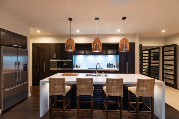 A sophisticated contemporary kitchen with dark timber cabinetry, integrated stainless-steel appliances, a large white island with four woven bar stools, and a bright blue-lit splashback. Three copper pendant lights hang above the island, while a glazed dark-framed door at right leads to an adjacent space.