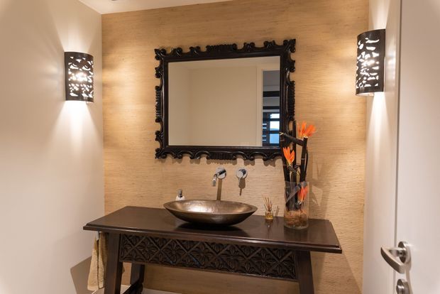 A compact, warmly lit bathroom vanity area featuring a dark stone benchtop, sculptural vessel basin, wall-mounted metal tapware, and an ornate black framed mirror. The rear wall has a textured tan finish, complemented by decorative black wall sconces and a carved dark vanity base.