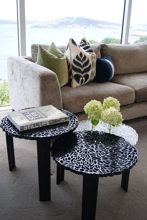 A bright, contemporary living room furnished with a large taupe sectional sofa, layered green, cream, black and patterned cushions, and two sculptural black side tables with irregular cut-out tops. A glass coffee table holds pale green flowers, while full-height glazing provides expansive water and mountain views.