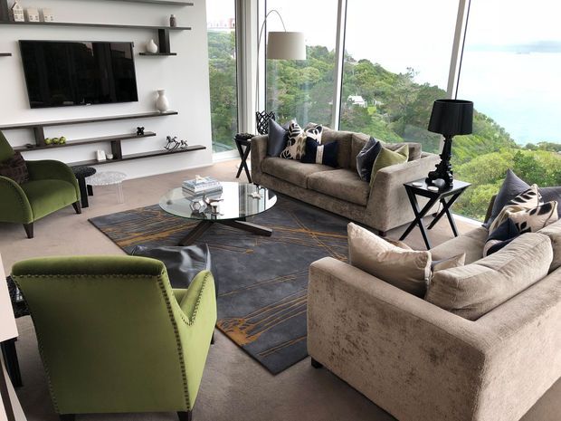 A bright, spacious living room with floor-to-ceiling corner glazing overlooking lush greenery and water. Two large neutral upholstered sofas and two olive-green armchairs surround a round glass coffee table on a dark patterned rug. Built-in open shelving and a wall-mounted television form a media wall, with black table lamps and monochrome cushions adding contrast.