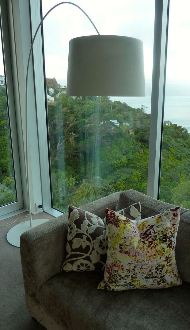 A contemporary living room corner with a large upholstered armchair or chaise positioned beside floor-to-ceiling corner glazing. A tall arched floor lamp with a large cylindrical shade rises over the seating, while patterned decorative cushions add colour and texture. The windows overlook a densely wooded, hilly residential landscape and water in the distance.