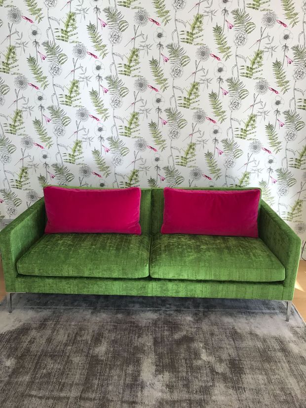 A compact green upholstered sofa with slim metal legs and two vivid magenta cushions sits against a light botanical-patterned wallpaper. A large distressed grey area rug covers the floor, creating a colourful, eclectic interior vignette.