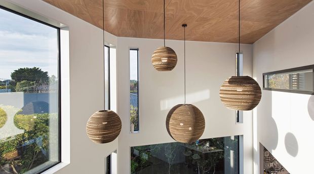 Double-height interior void with large floor-to-ceiling windows, white walls, a warm plywood-lined ceiling, and several suspended woven spherical pendant lights. The space feels bright and airy, with a glimpse of greenery and neighbouring buildings through the glazing.