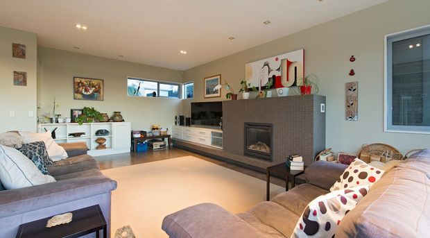 A spacious contemporary living room with a large L-shaped sectional sofa, media wall, and a prominent tiled fireplace surround. The room features pale grey walls, warm timber flooring, recessed ceiling lights, built-in white cabinetry, artwork, and a bright informal seating area by the window.