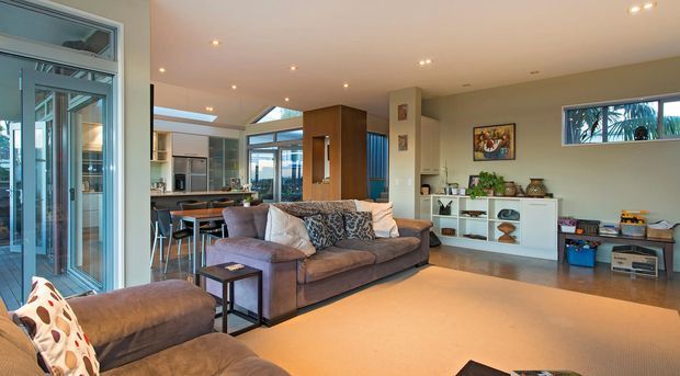 Open-plan contemporary living area with a large sectional sofa and ottoman in the foreground, facing a low white entertainment unit. The room connects directly to a modern kitchen and dining area, with extensive glazing and sliding doors providing abundant daylight. Neutral finishes, recessed ceiling lighting, timber cabinetry, and carefully arranged shelving create a warm, relaxed family interior.