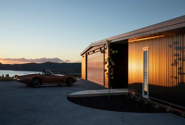 Exterior view of a sleek contemporary single-storey residence or garage volume at dusk, featuring a flat or very low-pitched roof, warm vertical metal cladding, a large timber-toned sectional garage door, and a narrow vertical window. A paved driveway and curved dark landscaped bed sit in front, with a vintage sports car parked beside the building. Water and rolling hills are visible in the distance.