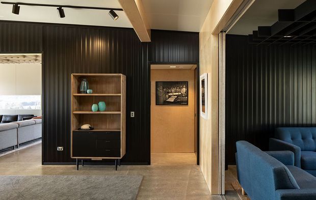 Interior view of a refined contemporary media or living space with dramatic black vertical wall panelling, warm timber door and shelving, polished concrete flooring, and blue upholstered seating visible in the adjoining room. A large opening connects to a brighter lounge area, while track lighting and a dark ceiling grid add a cinematic feel.
