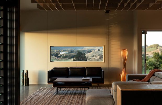 A warm, contemporary living room with a black leather sofa, low dark coffee table, textured area rug, and a large upholstered sectional or ottoman in the foreground. A sculptural floor lamp stands beside the seating, while broad windows and a horizontal picture window bring in views of the surrounding landscape. The room has a dark exposed grid ceiling, smooth warm beige walls, and a calm, refined atmosphere.