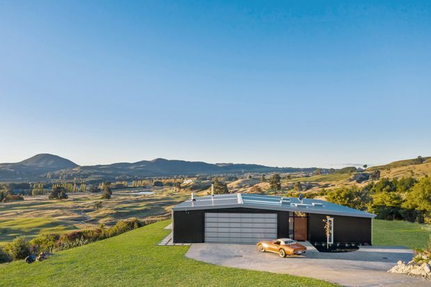 A single-storey contemporary dark-clad building set on a gently sloping rural property, with a low pitched metal roof, broad garage door, and covered entry or carport-like recess. The structure sits in a landscaped lawn overlooking an expansive agricultural valley and rolling hills under a clear blue sky. An orange car is parked in front of the building.
