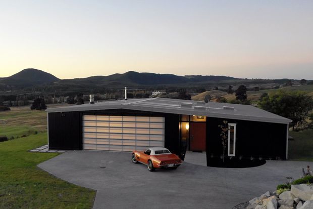 Exterior view of a low, contemporary rural home or garage-like residence with a broad flat roof, dark vertical cladding, and a large horizontal-panel glazed or sectional garage door. A paved forecourt and orange vintage car sit in front, with rolling countryside and hills beyond.