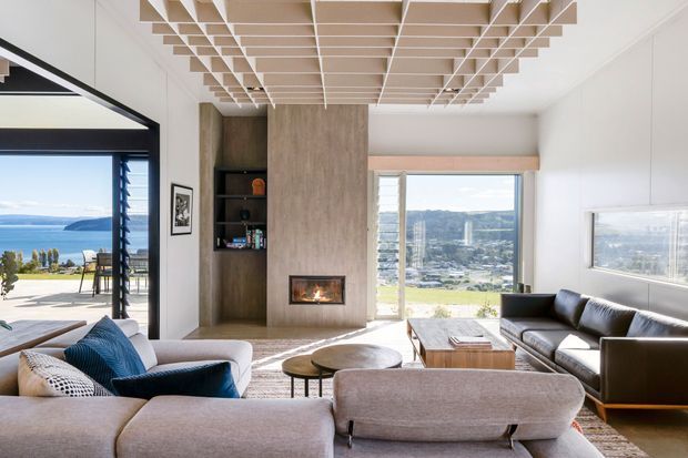 Bright contemporary living room with expansive glazing and views toward water and surrounding hills. The room features a large sectional sofa, leather sofa, timber coffee tables, a central stone or concrete fireplace surround with an inset firebox, built-in dark timber shelving, and a distinctive geometric timber ceiling grid. Outdoor paving and landscape are visible through the open glazing.