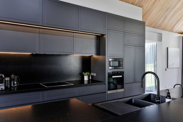A refined contemporary kitchen with extensive dark grey built-in cabinetry, integrated appliances, a black stone-look benchtop and matching splashback, and a large island with double sink and curved black tapware. Warm under-cabinet lighting contrasts with the pale timber-lined sloped ceiling and white walls. Glazing and artwork are visible beyond the kitchen.