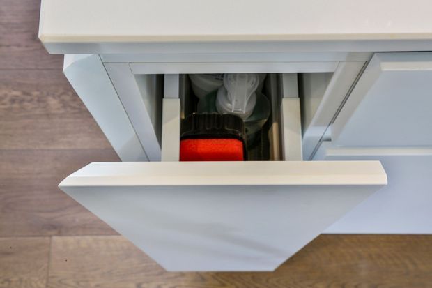Close-up, top-down view of a white built-in drawer opened to reveal stored kitchen items, including a red container and dishware. The cabinetry has recessed panel detailing and sits above a warm medium-brown timber-look floor.
