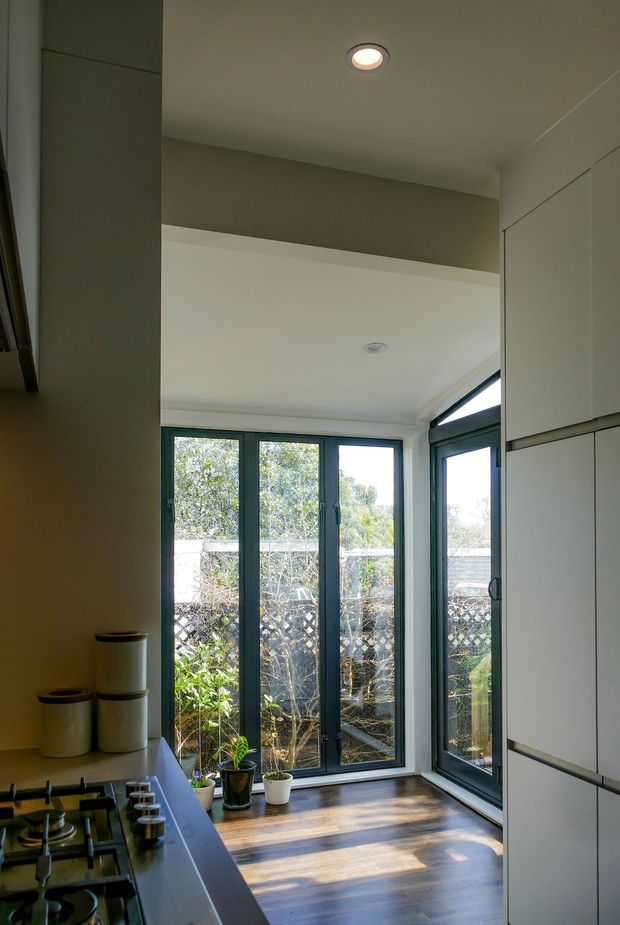 Compact contemporary kitchen with dark built-in cabinetry, a gas cooktop and stainless-steel controls in the foreground, opening to a bright glazed corner with tall dark-framed windows and a glass door overlooking a small planted outdoor area. Warm timber flooring, pale walls, recessed ceiling lights and integrated white cabinetry create a clean, light-filled space.