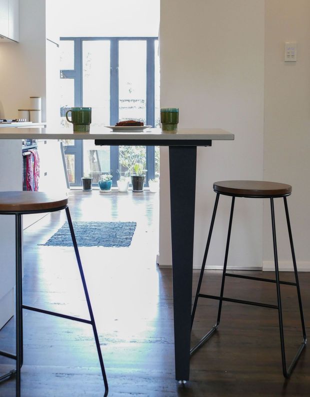 Bright, compact kitchen or dining nook featuring a slim island or breakfast bar with a light stone-like top and dark pedestal base. Two tall stools with round dark timber seats frame the island. The space has dark timber flooring, pale walls and cabinetry, and a large blue-grey framed glazed door or window opening to a light-filled area with potted plants. Green mugs and a plate sit on the island, adding a small accent of colour.