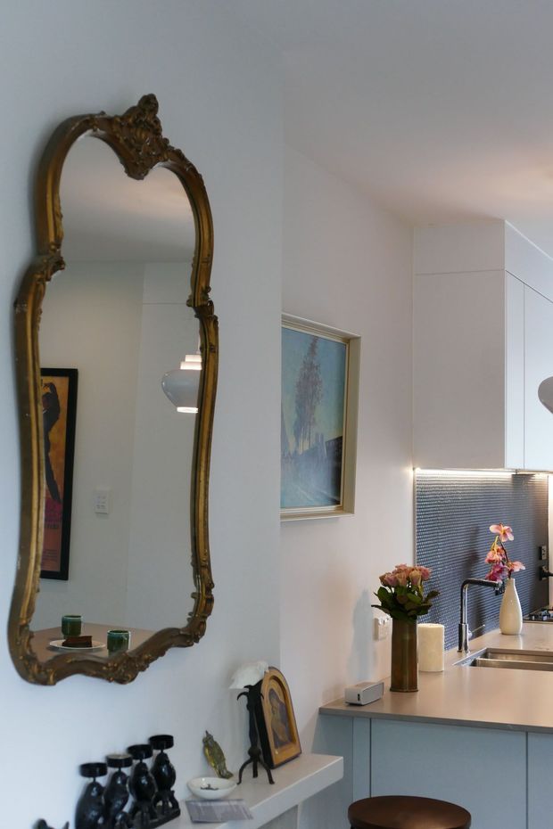 A refined contemporary kitchen viewed from an adjacent interior space, with pale blue-grey cabinetry, a light stone or engineered-stone benchtop, stainless-steel sink and tapware, and a textured metallic splashback illuminated by under-cabinet lighting. The white walls feature a large ornate antique-style arched mirror, framed artwork, and a narrow display shelf with decorative objects. A timber-toned stool and floral arrangements add warmth to the otherwise light, elegant space.