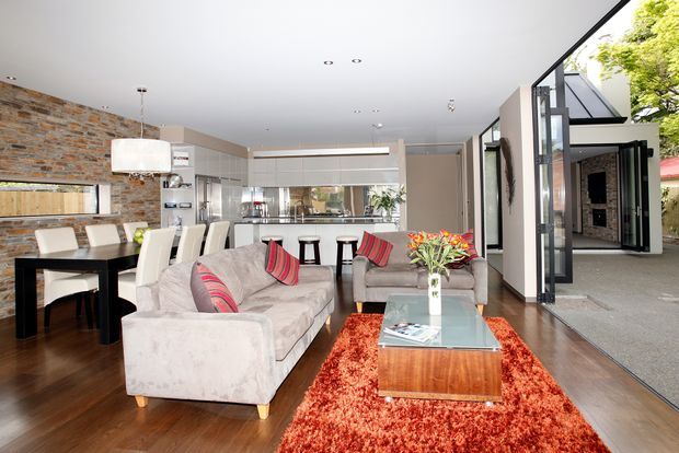 Open-plan contemporary living and dining area with a kitchen visible beyond. The room features a large sectional sofa, black dining table with upholstered chairs, glass coffee table, vivid red shag rug, exposed brick feature wall, timber flooring, extensive glazing, and a broad connection to an outdoor entry or courtyard area.