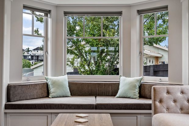 Bright built-in window seat in a traditional-style interior, with three large white-trimmed windows, roller blinds, a dark upholstered bench cushion and backrest, pale blue-green throw pillows, and white paneled cabinetry below. A tufted armchair and timber coffee table are partially visible in the foreground. Residential rooftops and leafy trees are visible outside.