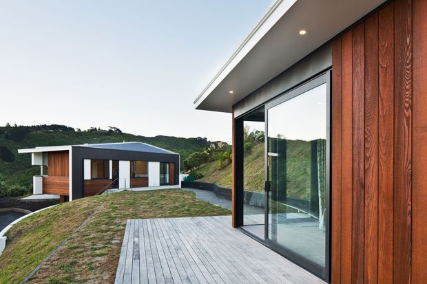 Modern residential exterior viewed from a timber-clad terrace or deck, with large black-framed sliding glass doors, a projecting flat roof and recessed downlights in the foreground. A separate low-slung contemporary house with dark roofing, timber wall sections and white rendered elements sits across a landscaped sloping site surrounded by dense greenery.
