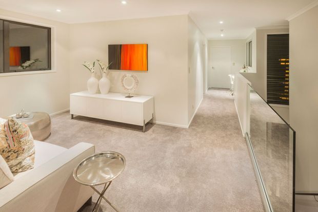 Bright, refined open-plan living and hallway interior with soft beige carpeting, white walls and built-in cabinetry. A white console with decorative objects sits beneath a vivid orange-red abstract artwork, while a pale sofa, patterned cushions and a glass side table occupy the foreground. The hallway extends toward a closed door and includes recessed lighting, a mirrored or glossy feature surface, and dark horizontal slatted cabinetry visible on the right.