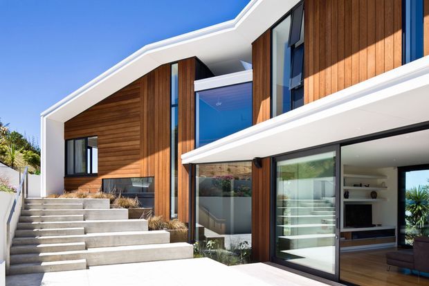 Exterior view of a contemporary multi-level home with strong geometric rooflines, white rendered framing, warm vertical timber cladding, extensive dark-framed glazing, and broad concrete steps leading to the entrance. A living room is visible through the ground-floor sliding doors.
