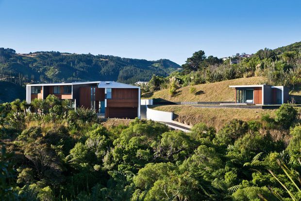 A pair of low-profile contemporary buildings integrated into a steep, densely vegetated hillside. The structures feature dark timber cladding, broad glazing, crisp white rendered walls, and shallow metal roofs, with a winding access path and dramatic forested hills beyond.