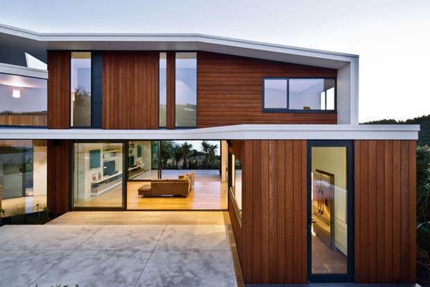 Modern two-storey residential house with a strong horizontal composition, flat rooflines, warm vertical timber cladding, dark-framed glazing, and a concrete-paved entry courtyard. Large glazed openings reveal a contemporary living area inside, while white and grey structural bands create a crisp geometric facade.