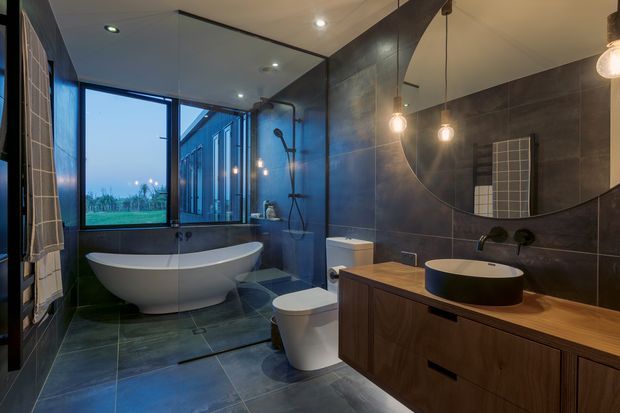 Contemporary bathroom with a freestanding white bathtub, walk-in glass shower, wall-hung toilet, vessel basin, and dark timber vanity. Large dark grey tiled surfaces create a moody, refined interior, complemented by a large round mirror and exposed pendant lights. Floor-to-ceiling glazing provides views to the exterior.