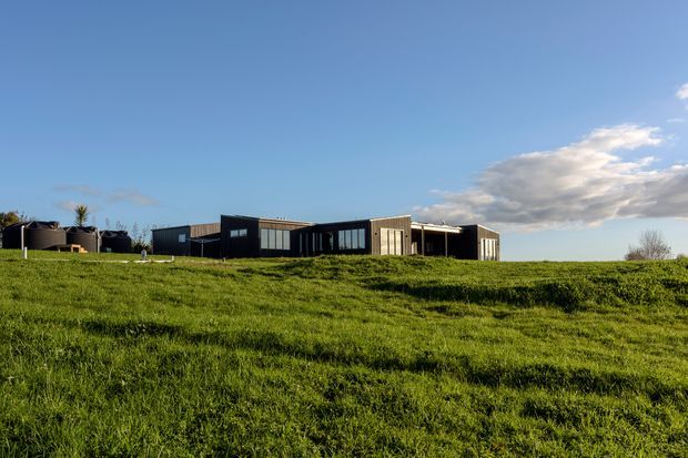 A low, elongated contemporary building set within an expansive grassy landscape. The dark facade features extensive horizontal glazing, vertical window bands, and a projecting flat roof, with a lighter timber-toned or pale panelled section near the centre. Large cylindrical tanks or utility structures sit to the left, while open green pasture and blue sky dominate the foreground and background.