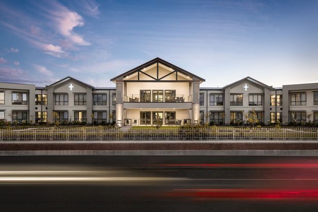 Symmetrical multi-storey institutional or commercial building viewed from across a busy roadway at dusk. The facade has a prominent central gabled entrance with exposed triangular framing, flanking gabled wings, repeated rectangular windows, bright interior lighting, and illuminated cross motifs. A landscaped forecourt and metal fence separate the building from the road, where passing traffic creates light trails.