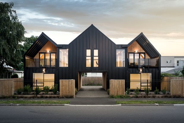 Front exterior view of a contemporary multi-unit residential building with three steeply pitched gabled forms, dark vertical metal cladding, large glazed openings, and timber privacy fencing. The central section forms a covered pedestrian passage, while the two outer units have balconies and warm illuminated interiors. The building sits along a suburban street at dusk.